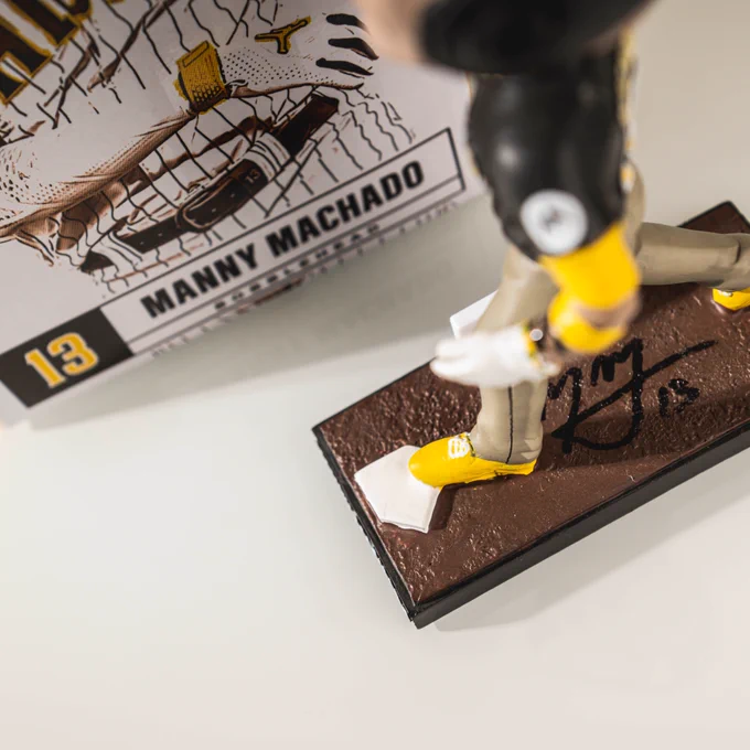 Additional photo of a Manny Machado bobblehead against a white backdrop. This photo also includes the box that the bobblehead comes in. The photo is being taken from above the box and bobblehead to show that Manny signed the base of the bobblehead.