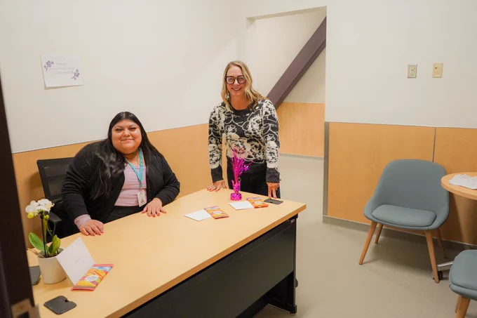 Two staff members pose for photos at a desk at the health center