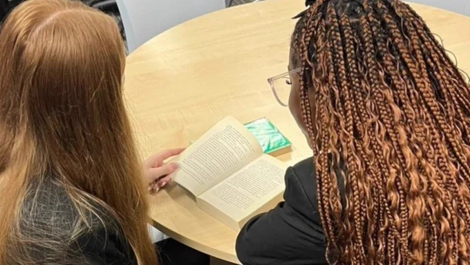 Two people sitting at a table, reading a book together in a library setting.