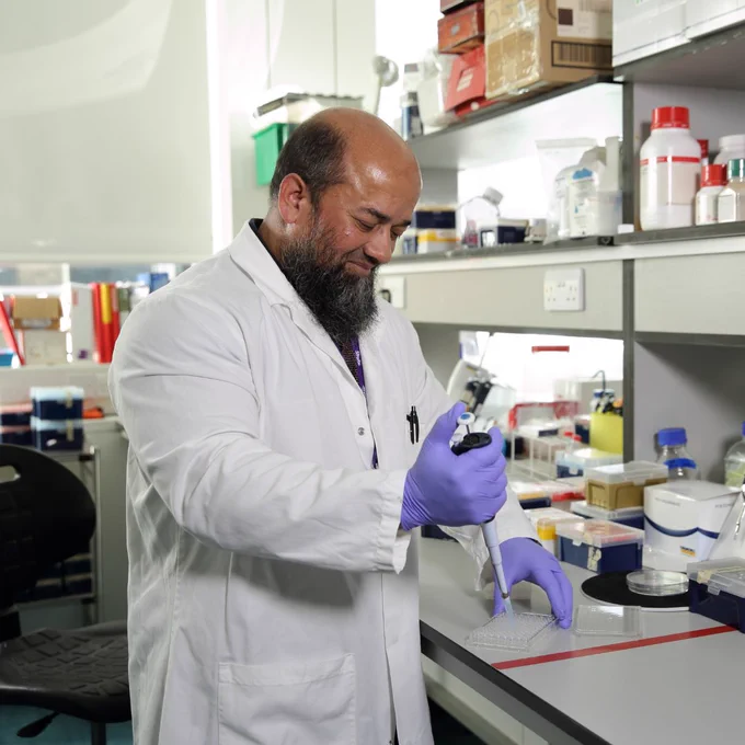 A man standing at a work bench with a pipette and vial. He's wearing a white lab coat. 