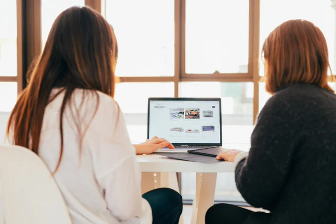 a photograph of two women sitting at a desk working on a laptop