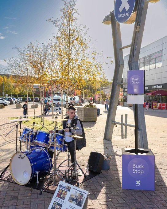 A musician is playing drums at a "Busk Stop" located outside the Xscape entertainment centre on a sunny day.