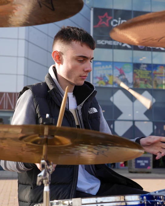 A person playing a drum set outdoors in front of the Xscape entertainment centre in Castleford, Yorkshire