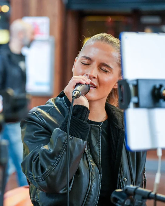 Person singing into a microphone, wearing a leather jacket, with recording equipment visible in the foreground at Xscape Yorkshire's Busk Stop.