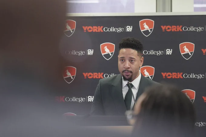 a man speaking in front of a york college backdrop