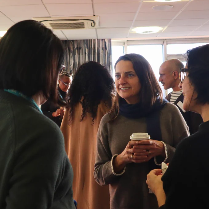Group of 3 people in discussion. Main person is holding a coffee cup and smiling. There are people in the background and they are in a lecture theatre.