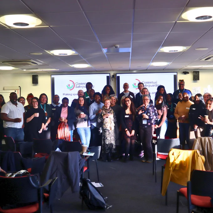 Group of people in front of two projection screens in a lecture theatre. People are smiling and have their arms around each other with coffee cups in their hands. 