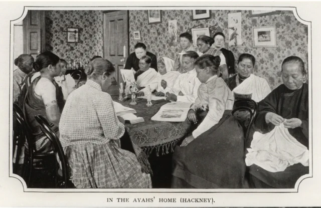 Black and white photo of the interior of the Ayahs’ Home in Hackney, with a number of women sitting around a table reading and embroidering.