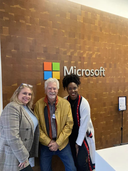 In order from left to right on image: Michele Erwin (founder), Blake Emery (board member), and Fey from Statuesque Events (AWU Global Forum planner) stand in front of a brown wall at Microsoft with the Microsoft logo on it.
