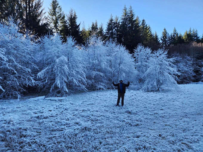 A man stands in a grass field that is covered in a layer of frost. He is about twenty feet away from the camera, looking towards it and raising both hands in the air. Behind him are a line of small trees also completely covered in frost. Behind these are taller trees that are being hit by the sun and have no ice on them.