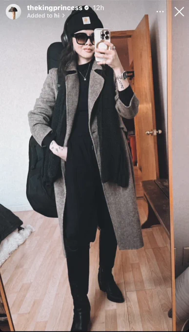 Ruibo Qian stands in front of a miror in a black outfit and brownover jacket with a black guitar case and black scarf taking a selfie