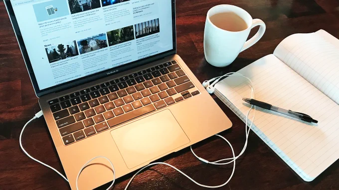 Horizontal photo of a laptop open on a wood grain desk, staged with headphone cords, an open notebook, pen, and mug. Original photo credit Alex Witze.