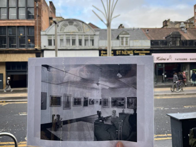 Looking over to New Charing Cross Gallery - the bank of windows with grey surround above a vacant shop. Holding the sheet that shows a photograph of the interior of the gallery (photograph held by National Library of Scotland), which depicts a wall of paintings and sculptures and a table in the foreground).