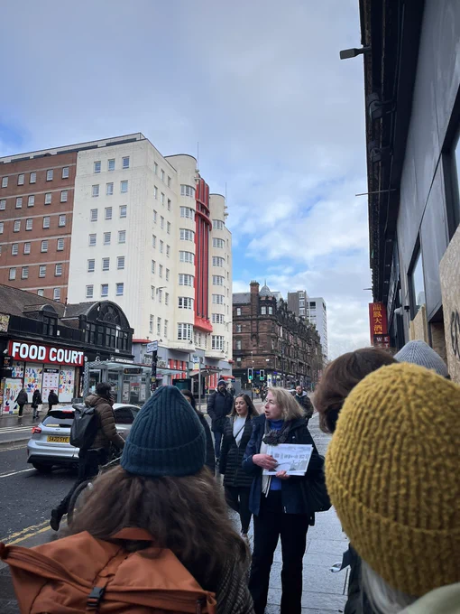 A group stands on Sauchiehall St learning about New Charing Cross Gallery that Bet Low co-founded in the 1960s. It’s a cold dry day so those on tour wearing woolly hats and warm coats.