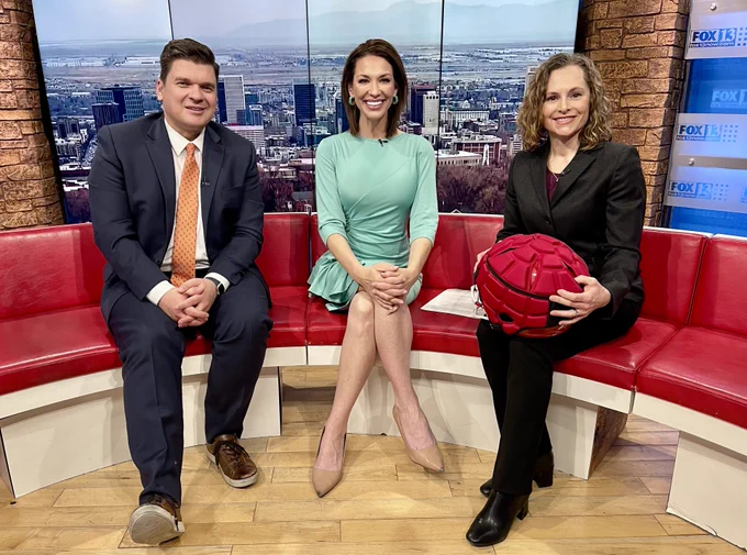FOX13 Good Day Utah anchors, John Franchi and Kelly Chapman, sit next to MountainStar Healthcare sports medicine physician, Dr. Andrea Matich, on set, smiling. Dr. Matich is holding a red football helmet with additional padding. She works at Peak Orthopaedics at Lone Peak Hospital in Draper, Utah.