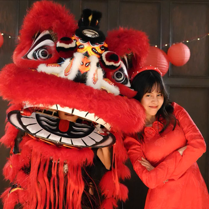 Person in a red dress standing next to a traditional Chinese lion dance costume, with red lanterns in the background.