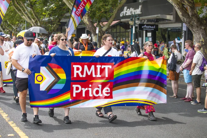 'RMIT Pride' banner with the inclusive flag rainbow pixel and rainbow design is proudly carried down the street by four RMIT community members
