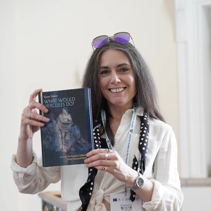 Susan Deacy holding a copy of her book *What Would Hercules Do? Lessons for Autistic Children Using Classical Myth* in front of a statue of Hercules on a neoclassical Warsaw staircase