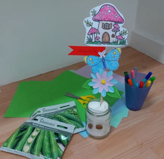 Photo showing a craft table surface with sheets of green, blue and pink coloured paper, a pot of colouring pens, a pair of scissors with yellow handles, two envelopes with dried peas and beans, and a glass jar. The glass jar has been filled with rolled up kitchen towel, with two dried beans wedged in-between the side of the jar and the paper. Stuck in-between the folds of the kitchen towel is a plant stick decorated with coloured-in cut-outs of a toad stool fairy home, a butterfly, a flower, two green leaves and a red banner.