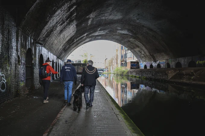 The Lord Mayor of Birmingham walks under a large bridge beside a canal, with Matthew Vaughan, Duncan the Civic Dog and a member of the River and Canal team in Birmingham.