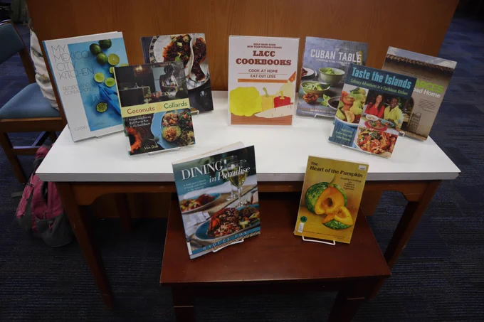 A display table showing Latin American & Caribbean cookbooks with a sign that says "Cook at Home / Eat out Less"