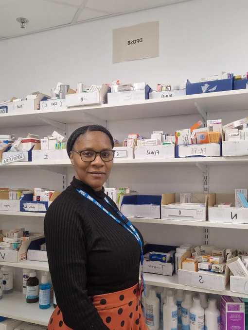 Sereita, a Pharmacy Technician Apprentice, stands in front of shelves filled with labeled boxes of medications and pharmaceutical supplies.