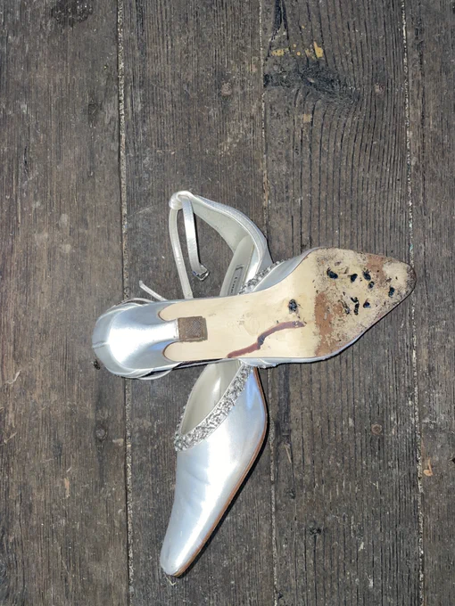 A pair of silver satin kitten heels lay on top of each other on a worn wood floor. The top shoe is flipped over to reveal a cluster of black flies crushed on the sole. Photo by Marie Ségolène Brault. 