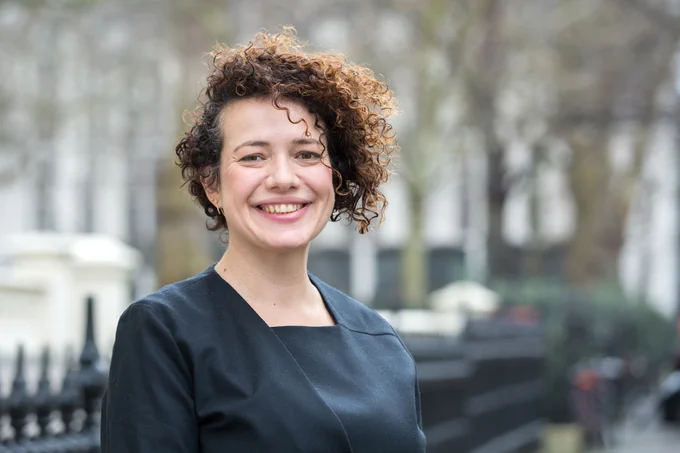 A portrait of Professor Marisa Miraldo with curly hair, wearing a black outfit, standing outdoors with trees and buildings in the background.