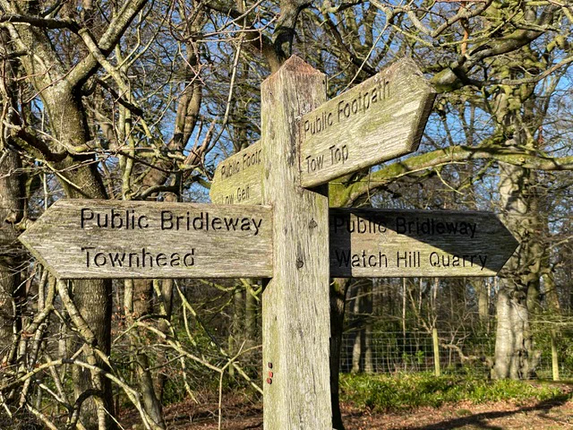Wooden signpost with multiple directions including "Public Bridleway Townhead," "Public Footpath Tow Top," and "Public Bridleway Watch Hill Quarry," set against a background of leafless trees and clear blue sky.