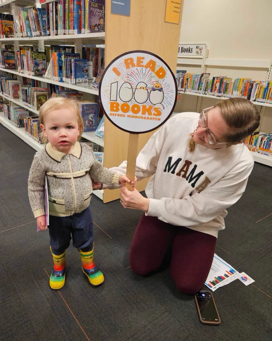 A young child and their caregiver pose with a sign that reads, " I read 1000 Books Before Kindergarten".