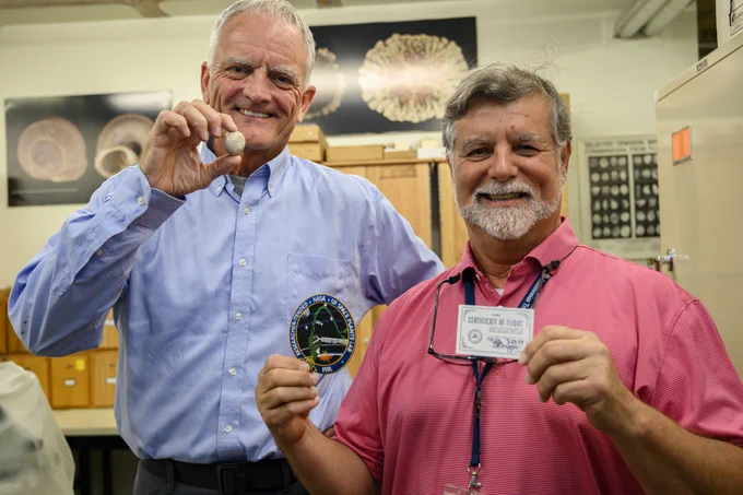 two older men are holding fossils and cards of some kind that are slightly blurry to read while standing for a portrait in a scientific fossil lab of some kind