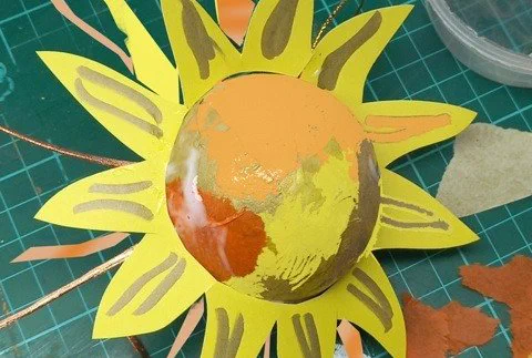 A piece of art made from a bauble, paint, paper and card to look like a sun.