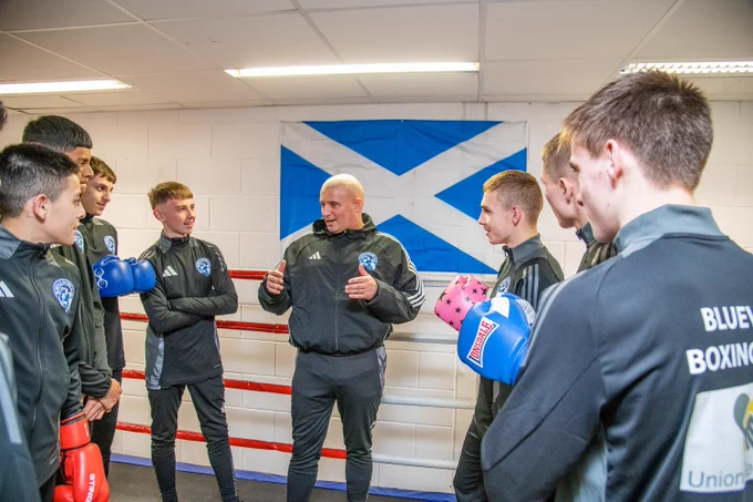 The pictures shows a boxing coach surrounded by male boxers all within a boxing ring. In the background is a Scotland flag and the boxers and coach are wearing tracksuits.