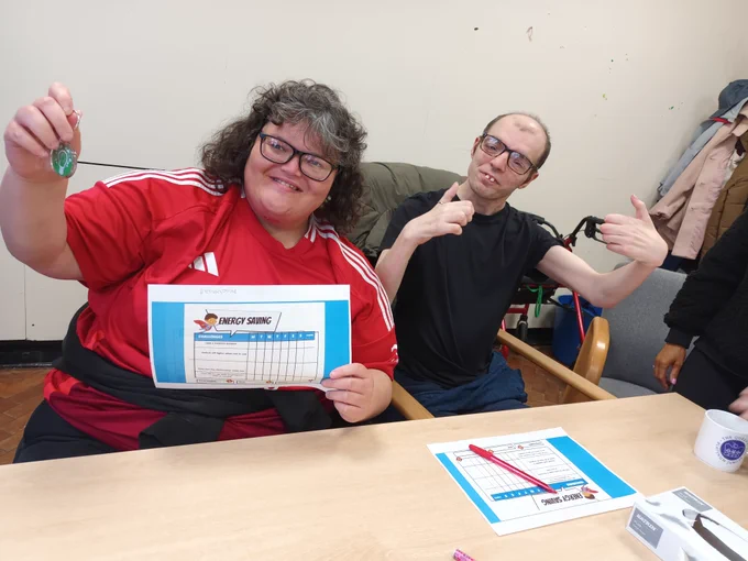 2 individuals smiling for the camera. One is holding up her energy saving worksheet and energy saver keyring.