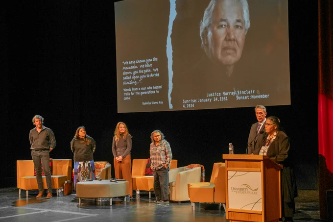 Guest speakers on-stage at the Mission Clarke Theatre, listening to Dr. Shirley Hardman from the University of the Fraser Valley at the podium