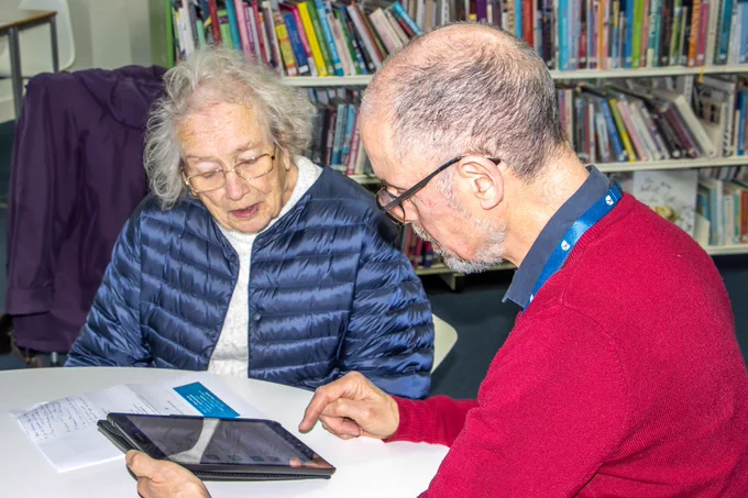 Photo of  man wearing a red jumper showing a lady in a blue coat how to use her tablet. They are sitting in a library setting.