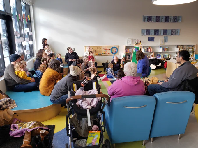 A white librarian reads a flannel board story to a group of children and their parents.
