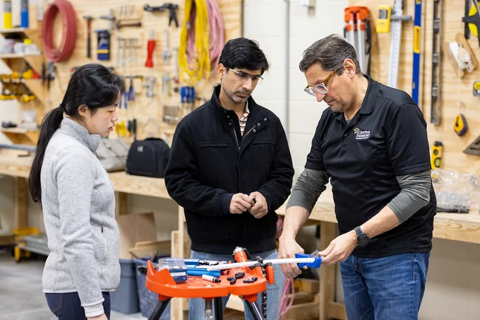 Three people engaged in a discussion around a workbench in a workshop, with one person demonstrating the use of a tool. The setting includes various tools and equipment in the background.