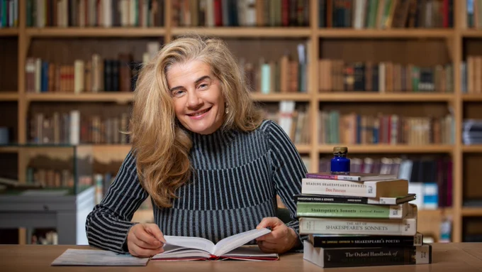 A photo of a woman in smart dress sitting at a desk with books, with shelves of books in the background
