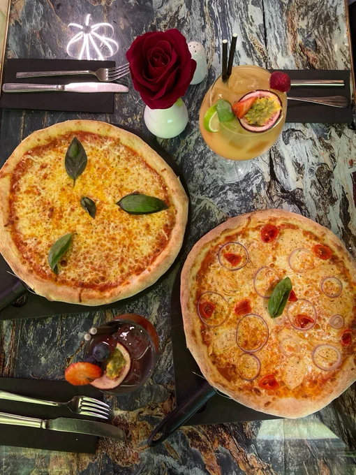 Two pizzas on a marble table, one with basil leaves and the other with tomato slices and basil. Accompanied by two cocktails and a red rose.