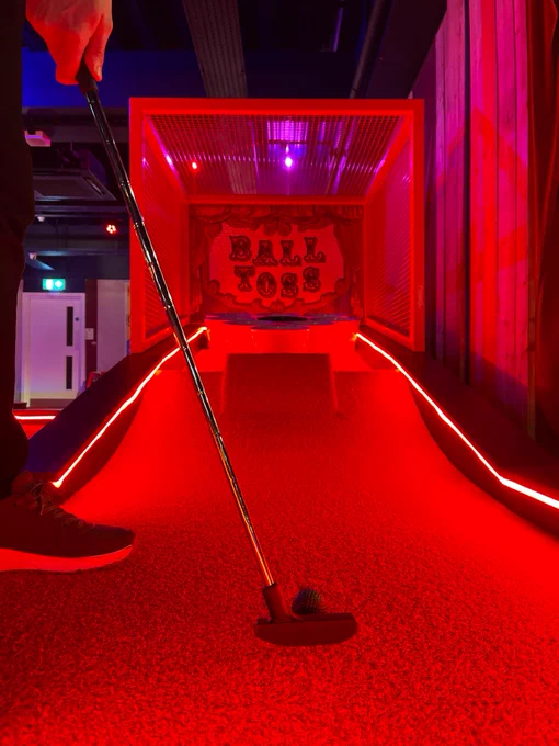 Person playing a carnival game called "Ball Toss" in a vibrant alley illuminated by red and purple lights.
