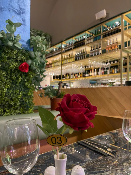 Red rose on a dining table at a restaurant with a shelf of wine bottles in the background.