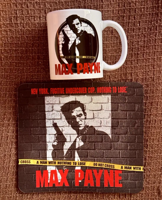 A Max Payne-themed coffee mug and mouse pad set displayed on a textured brown surface. The mug features the iconic noir-style image of Max Payne with a pistol against a brick wall, surrounded by yellow “DO NOT CROSS” tape. The mouse pad matches the mug’s aesthetic, with the same black-and-white image of Max Payne, the game’s red logo, and the tagline: “New York. Fugitive Undercover Cop. Nothing to Lose.” A nostalgic tribute to the classic 2001 third-person shooter.