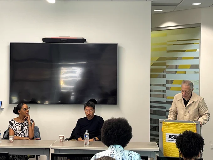 Medium shot of Lisa Lucas and Kirby Kim sitting together at a table in front of an audience. Ron Broglio speaks at a podium to the right.