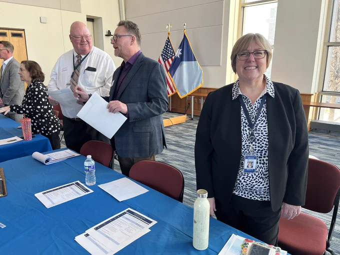 A woman smiles at the camera while two men discuss procurement at the MNIT IT Vendor Fair.
