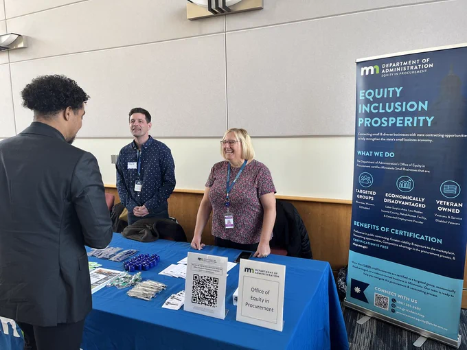 A woman discusses procurement with a vendor at her table during the MNIT IT Vendor Fair. The sign behind her reads, "Equity, Inclusion, Prosperity."