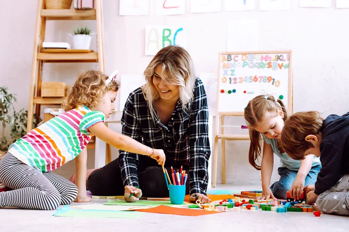 Three children and an adult participate in an arts and crafts activity on the floor, surrounded by colorful papers and craft supplies, in a bright, playful room with educational decorations on the walls.