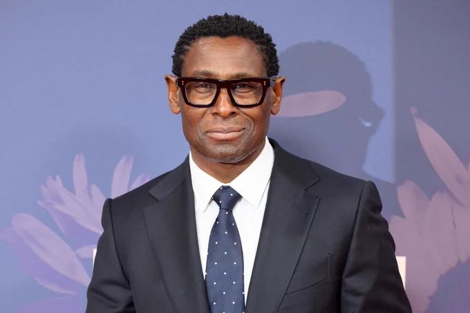 David Harewood, wearing glasses and a dark suit, attends a formal event with a floral background.