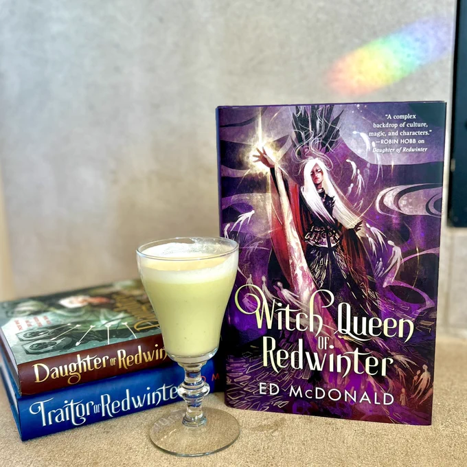 [image desc: A pale green cocktail sits alongside WITCH QUEEN OF REDWINTER by Ed McDonald. Behind the glass is a copy of DAUGHTER OF REDWINTER and TRAITOR OF REDWINTER by Ed McDonald. A rainbow flashes across the wall behind.]