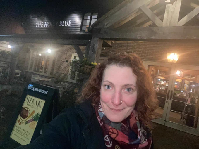 Stacy, a white woman with red curly hair in a scarf with peace symbols on it and a blue coat, stands smilng outside a warm, inviting looking pub called The Holly Blue